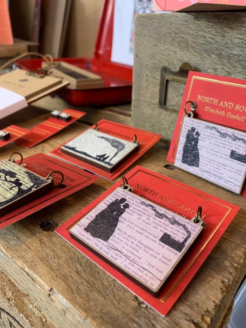 A photo of a series of classic literature theme jewellery pieced by Loadofolbobbins. Laser cut and showing silhouette scenes inspired by the novels North and South, Wuthering Heights and Alice in Wonderland. The silhouette scenes are shown against the original text from the novels.