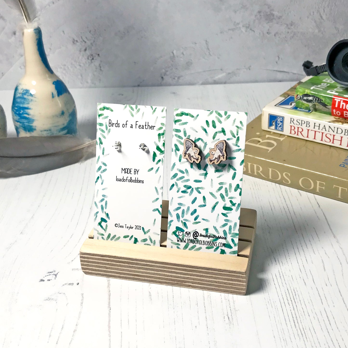 A pair of laser cut wooden earrings showing an illustration of a swooping barn owl bird by loadofolbobbins. The earrings are mounted on a mini backing card with a repeat abstract watercolour leaf pattern. They are standing in a wooden stand and in the background you can see a pile of bird watching books and a pair of binoculars.