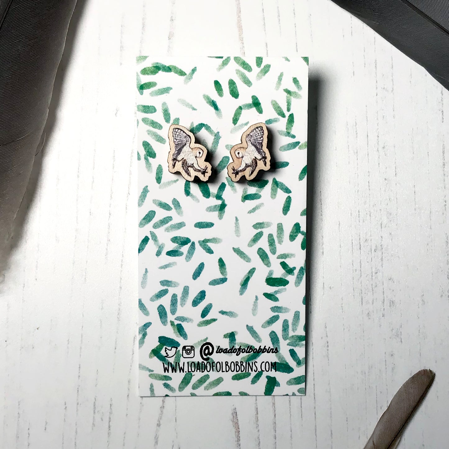 A pair of laser cut wooden earrings showing an illustration by Loadofolbobbins of a flying barn owl. The earrings are mounted on a mini backing card with a repeat abstract watercolour leaf pattern.