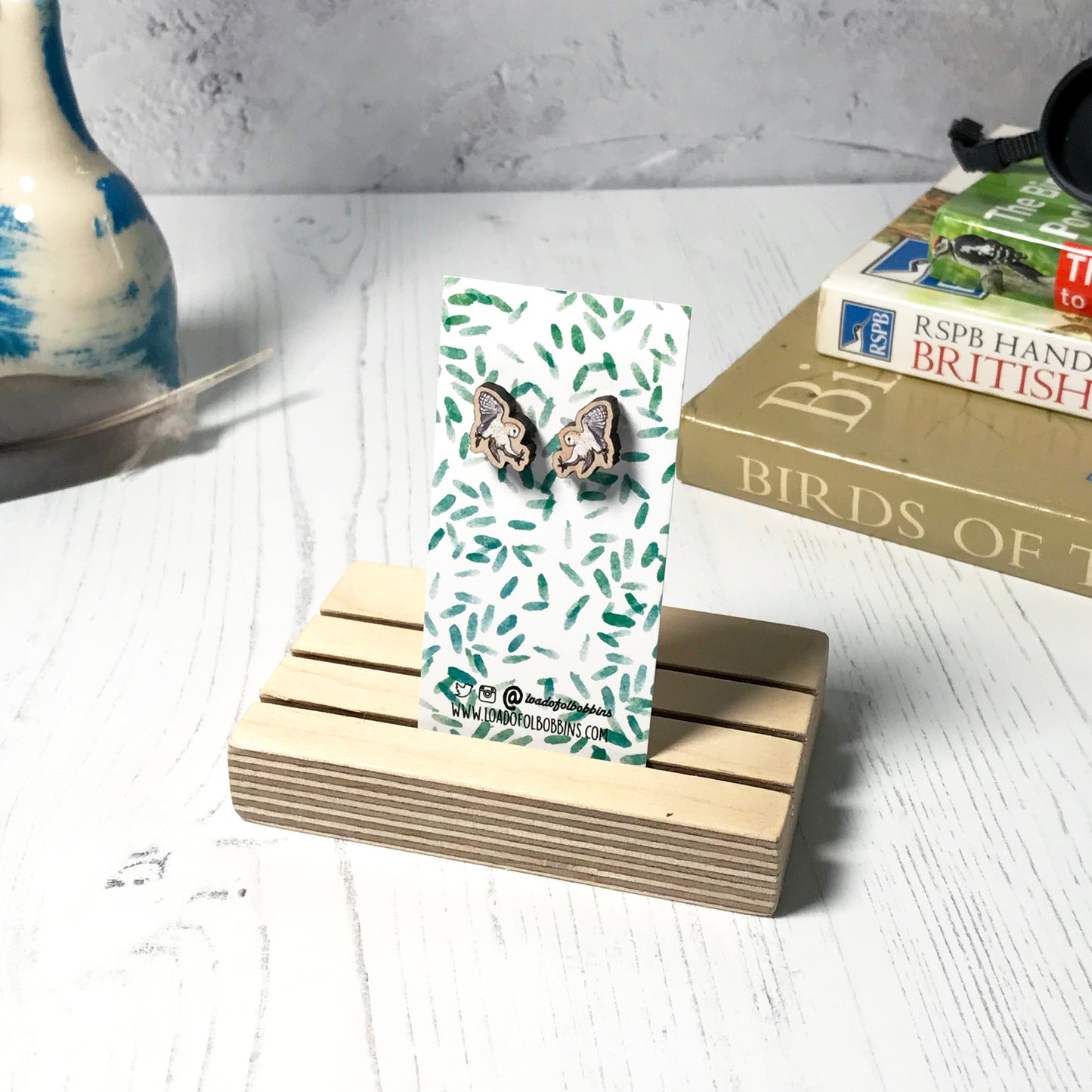 A pair of laser cut wooden earrings showing an illustration of a swooping barn owl bird by loadofolbobbins. The earrings are mounted on a mini backing card with a repeat abstract watercolour leaf pattern. They are standing in a wooden stand and in the background you can see a pile of bird watching books and a pair of binoculars.