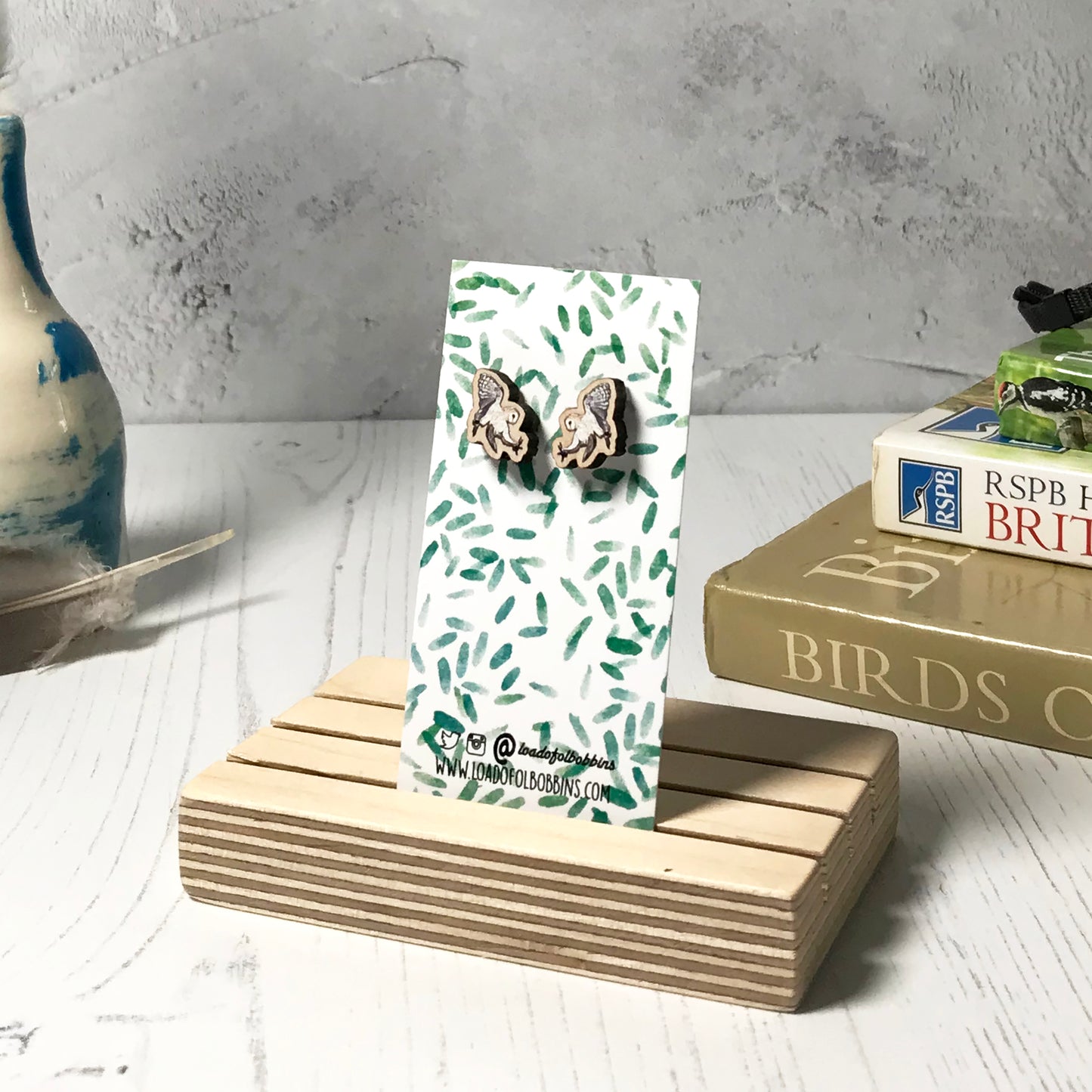 A pair of laser cut wooden earrings showing an illustration of a flying barn owl bird by loadofolbobbins. The earrings are mounted on a mini backing card with a repeat abstract watercolour leaf pattern. They are standing in a wooden stand and in the background you can see a pile of bird watching books and a pair of binoculars.