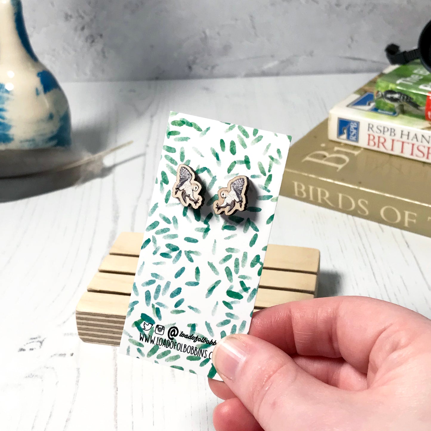 A pair of laser cut wooden earrings showing an illustration of a swooping barn owl bird by loadofolbobbins. The earrings are mounted on a mini backing card with a repeat abstract watercolour leaf pattern. They are being held by a hand for scale, which shows that each stud earring is slightly smaller than a thumbnail.