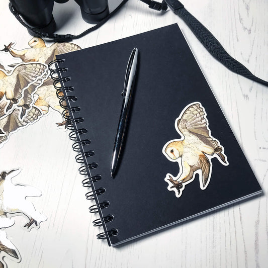 A photo of an eco-friendly glossy sticker about 8cm in height and width. The sticker is of original digital illustration by Loadofolbobbins showing a Barn Owl in flight and is stuck to the cover of a black spiral bound notebook with a pen next to it. The owl is swooping with claws beneath it to catch prey. It’s wings are together behind it caught in the upward flapping motion. The colouring of the owl is a combination of creams and browns with dark speckled markings across the body, underwing and head.