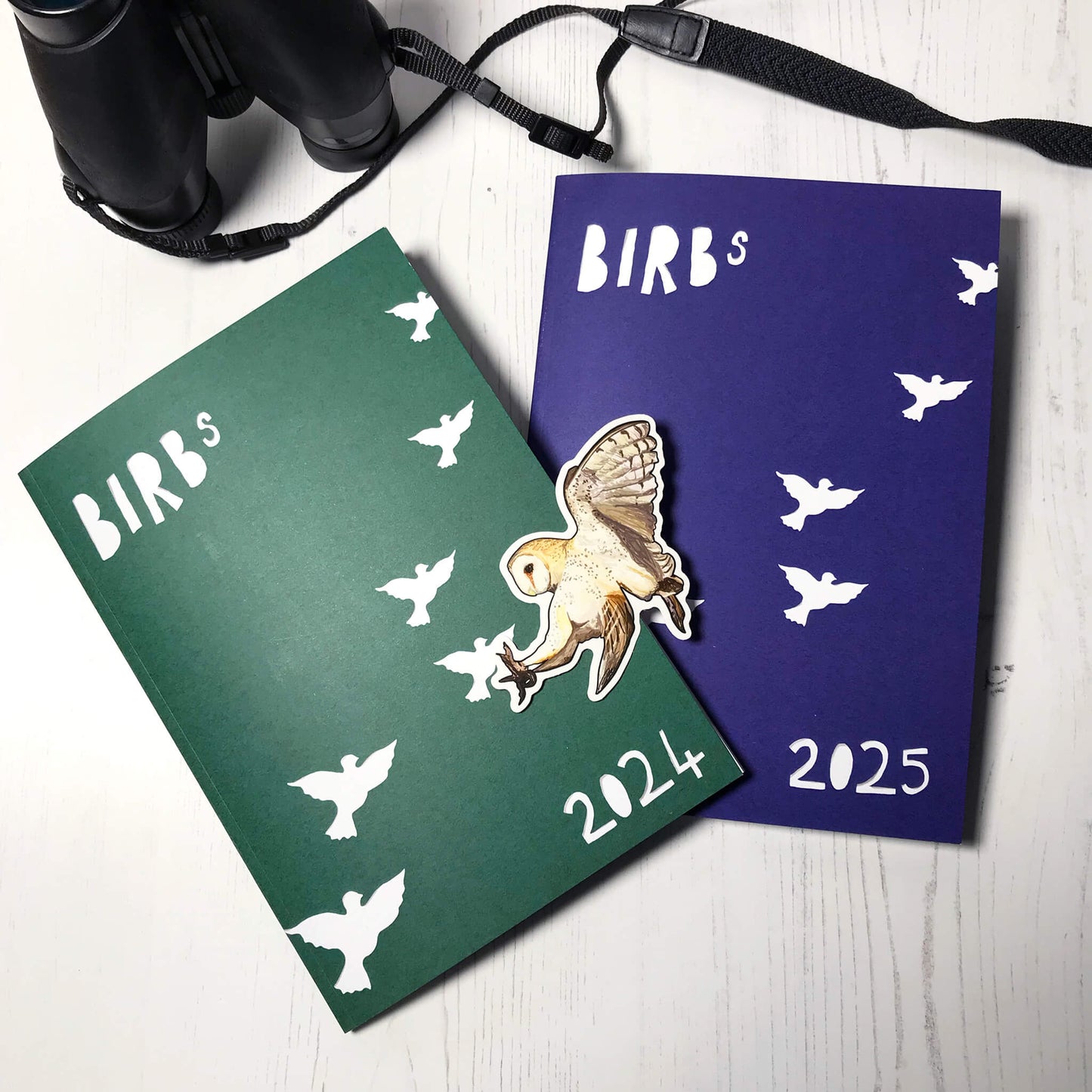 A photo of the covers of two printed zines full of bird illustrations by Loadofolbobbins. One on the left has a green cover with text Birbs 2024 and silhouettes of bird flying across it all in paper cut style. The zine on the right has a blue cover with the same design but 2025 instead. There's also a 8cm glossy sticker showing an illustration of a barn owl in flight, swooping with it's legs out beneath it as if about to catch some prey.