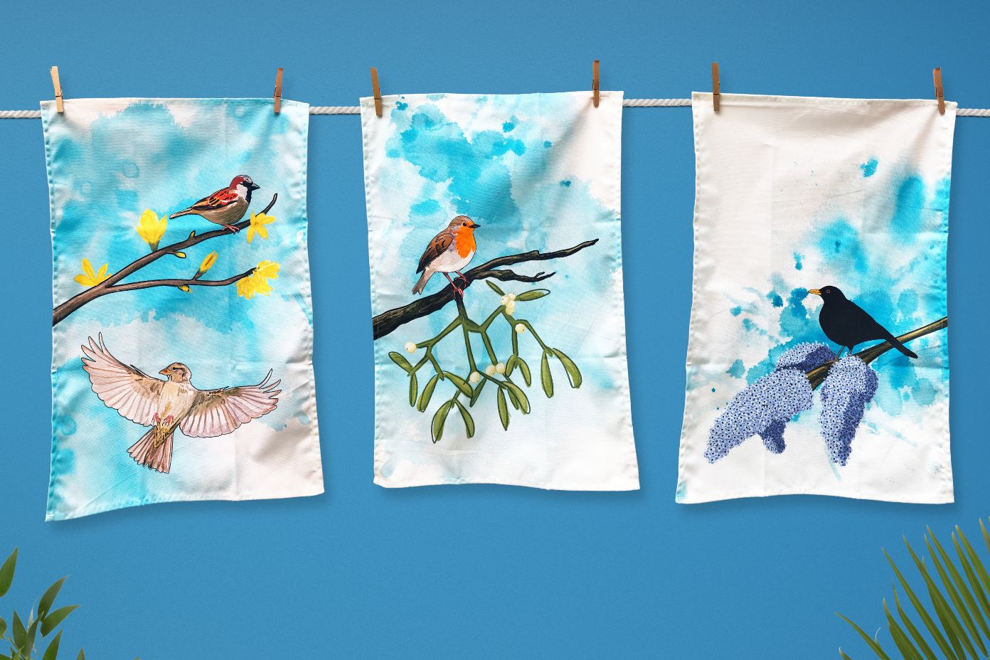 Three tea towels hanging on a washing line. Created from illustrations by Loadofolbobbins. From left to right they show a male sparrow perched on a forsythia branch with yellow flower blooms, beneath this is a flying female sparrow with wings spread out, a robin perched on a branch of mistletoe and a male black bird perched on a branch of lilac with three full purple blooms on the end. The backgrounds are all an inky wash in bright turquoise.