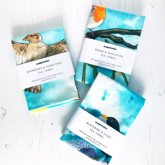 A collection of bird themed illustration tea towels by loadofolbobbins. Three folded tea towels one shows a female sparrow flying, a robin perched on a branch of mistletoe and a male blackbird on blooming lilac branch.