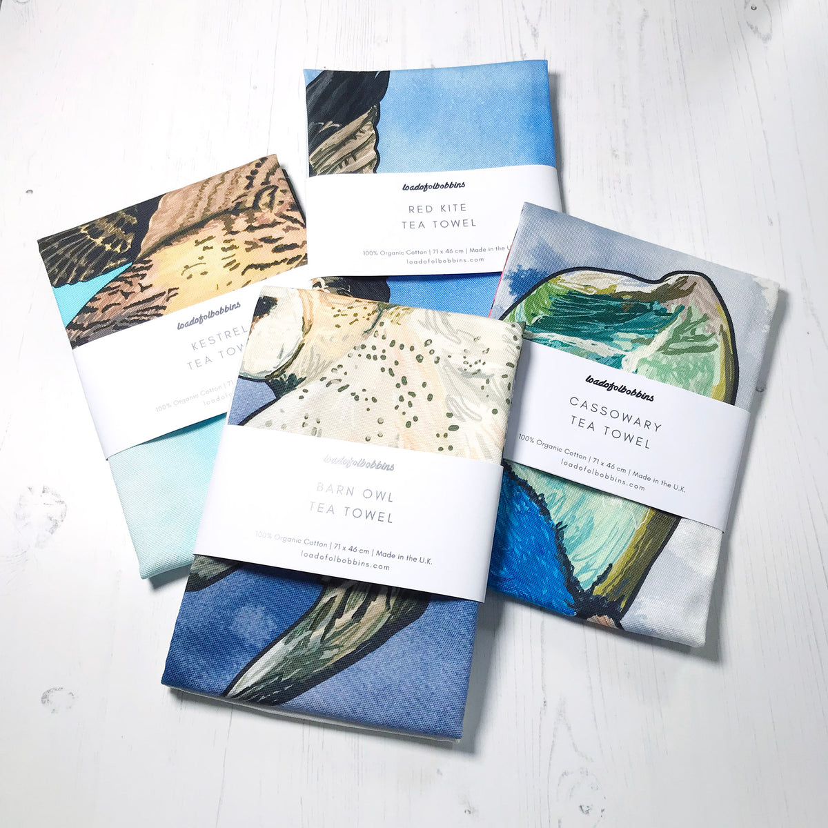 A collection of bird themed illustration tea towels by loadofolbobbins. Four folded tea towels one shows a hovering kestrel, a swooping barn owl, the head of a cassowary bird and a lying red kite.