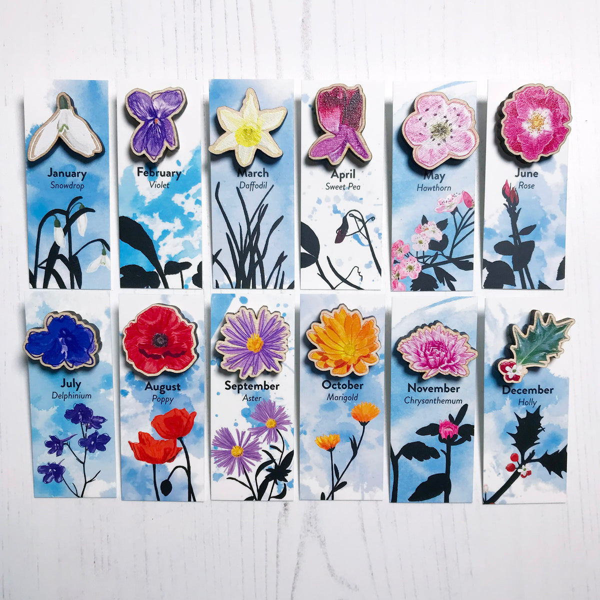 A collection of twelve flower themed wooden mini pins, mounted on illustrated backing cards, representing the birth flower for each month of the year. They are white snowdrop, purple violet, yellow daffodil, purple sweet pea, pinky white hawthorn, dog rose, blue delphinium, red poppy, purple aster, orange marigold, pink chrysanthemum and green and red holly.