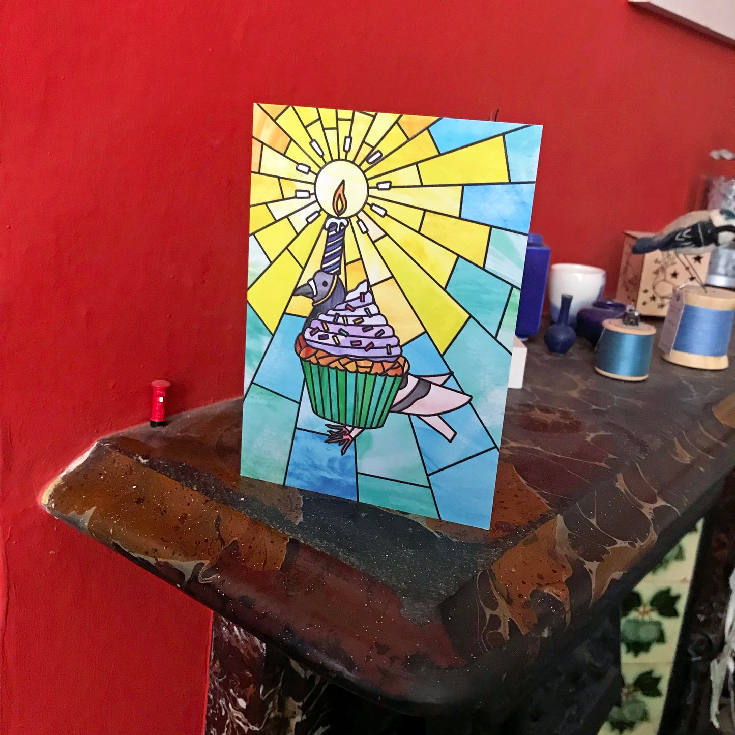 A photo of a greetings card displayed on a mantelpiece featuring an original illustration by Loadofolbobbins. The illustration shows a pigeon dressed up as a birthday cupcake with purple icing and rainbow sprinkles. Strapped to the pigeon's head is a lit birthday candle with light rays coming off the flame. The illustration is done in a stained glass style.