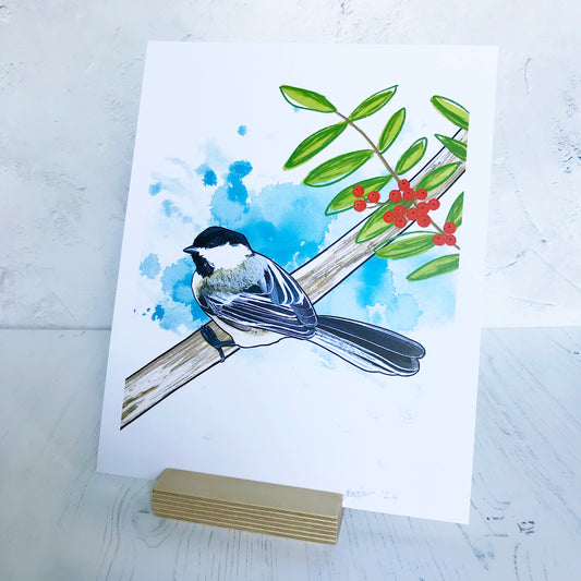 A print of an original digital illustration by Loadofofolbobbins of a black capped chickadee perching on a branch next to some bright red rowan berries and vibrant green leaves. A small tit bird with a fairly long tail with grey blue feathers. It has a black cap on its head with white cheeks and black under the beak. It's back and wings are also a grey blue colour. The background of the illustration is a series of light blue inky splashes. The bird is looking out to the left of the image.