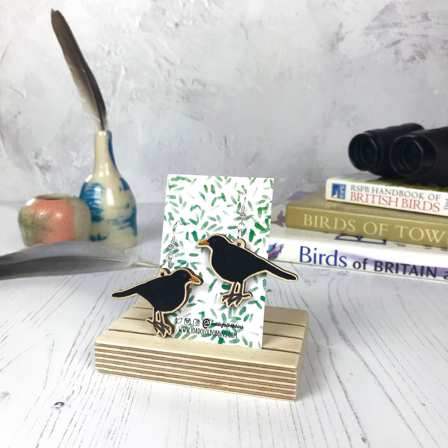 A pair of laser cut wooden dangle earrings showing an illustration of a male blackbird by loadofolbobbins. The earrings are mounted on a backing card with a repeat abstract watercolour leaf pattern. They are standing in a wooden stand and in the background you can see a pile of bird watching books and a pair of binoculars.
