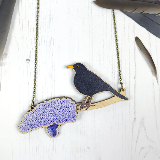 A laser cut wooden charm necklace showing an illustration of a male blackbird perching on a branch with two purple lilac blossoming blooms by loadofolbobbins. One of the main blooms stands proud of the necklace in a 3D relief effect.  The wooden necklace is hanging from an antique bronze cable chain.