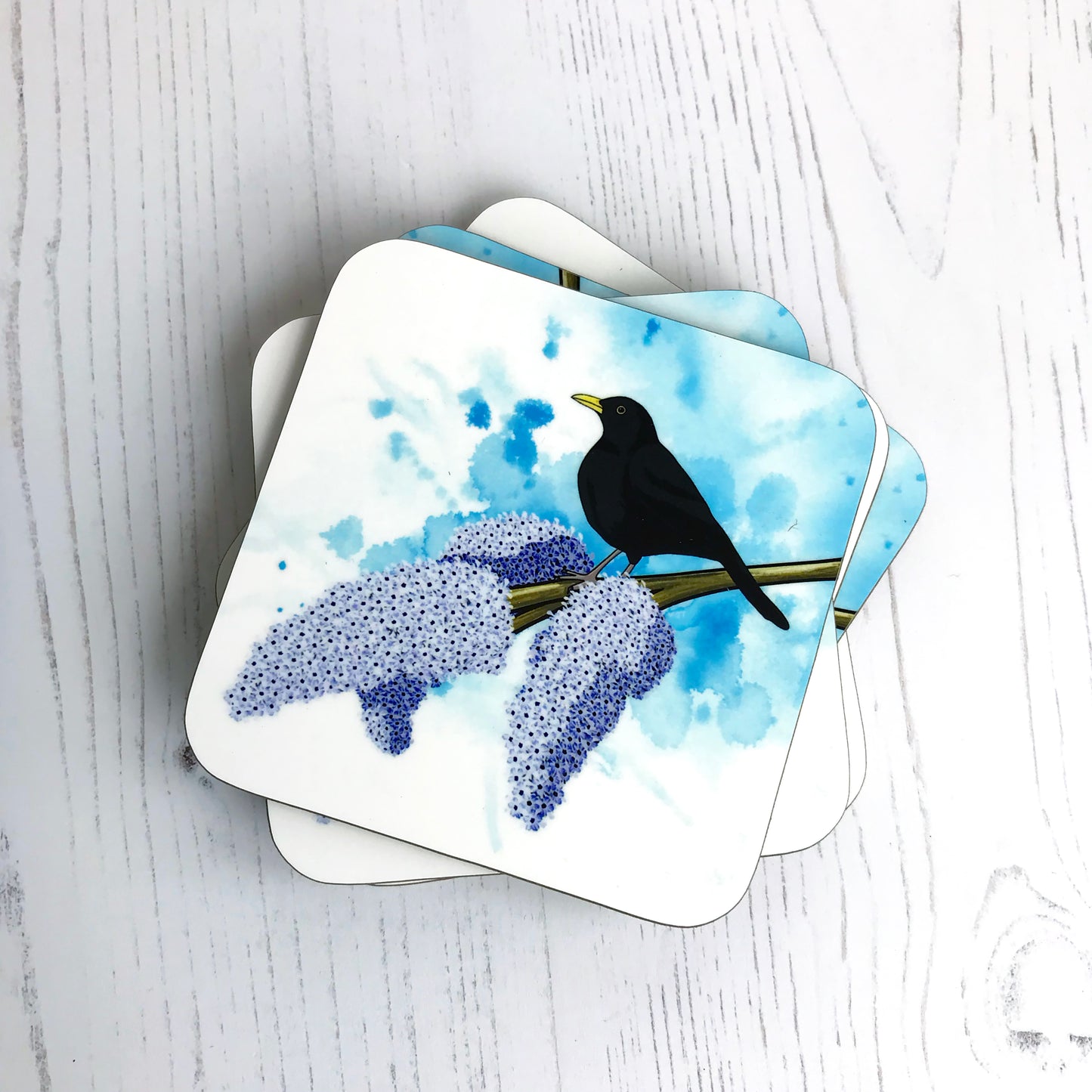 A photo of a pile of round edged square coasters which have an original illustration by Loadofolbobbins showing a male black bird perched on a branch of lilac with three full purple blooms on the end. The background is an inky wash in bright turquoise.