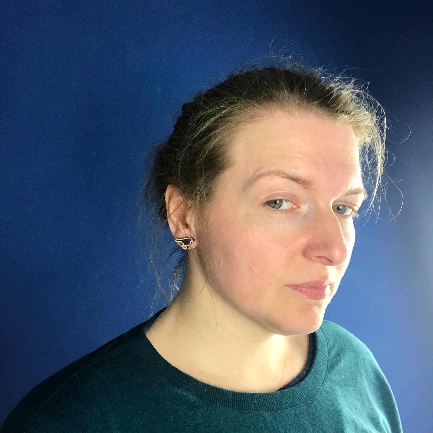 A neck and head close up photo of a white woman with blondish brown hair worn up and standing against a dark blue wall. Her face is slightly turned away from the camera so the main focus of the image is her ear which is wearing a small laser cut wooden stud earring of a male blackbird by Loadofolbobbins.