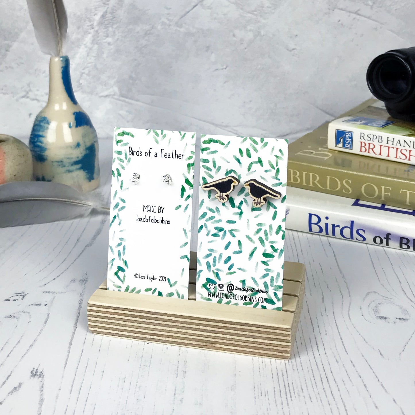 A pair of laser cut wooden earrings showing an illustration of a male blackbird by loadofolbobbins. The earrings are mounted on a mini backing card with a repeat abstract watercolour leaf pattern. They are standing in a wooden stand and in the background you can see a pile of bird watching books and a pair of binoculars.