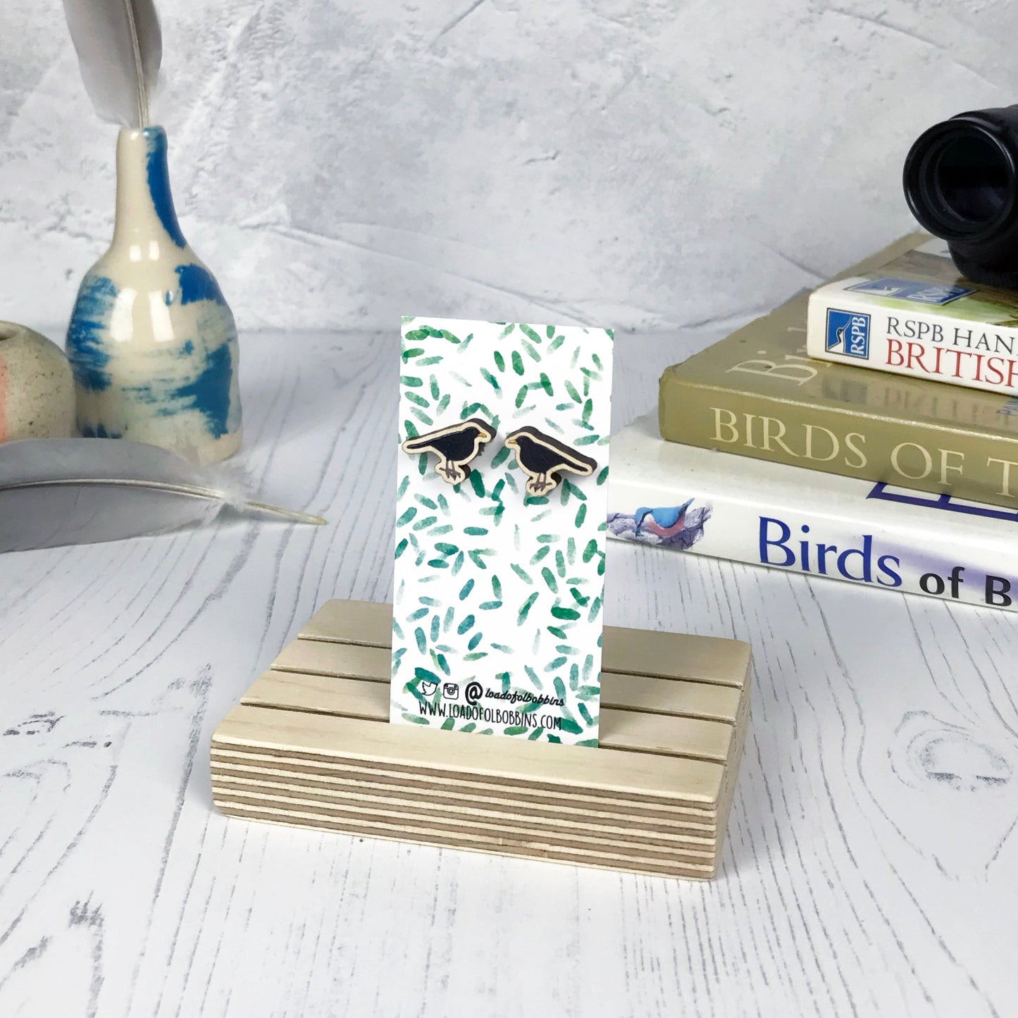 A pair of laser cut wooden earrings showing an illustration of a male blackbird bird by loadofolbobbins. The earrings are mounted on a mini backing card with a repeat abstract watercolour leaf pattern. They are standing in a wooden stand and in the background you can see a pile of bird watching books and a pair of binoculars.