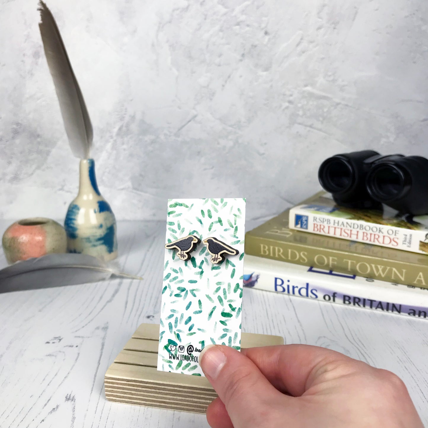 A pair of laser cut wooden earrings showing an illustration of a male blackbird by loadofolbobbins. The earrings are mounted on a mini backing card with a repeat abstract watercolour leaf pattern. They are being held by a hand for scale, which shows that each stud earring is slightly smaller than a thumbnail.