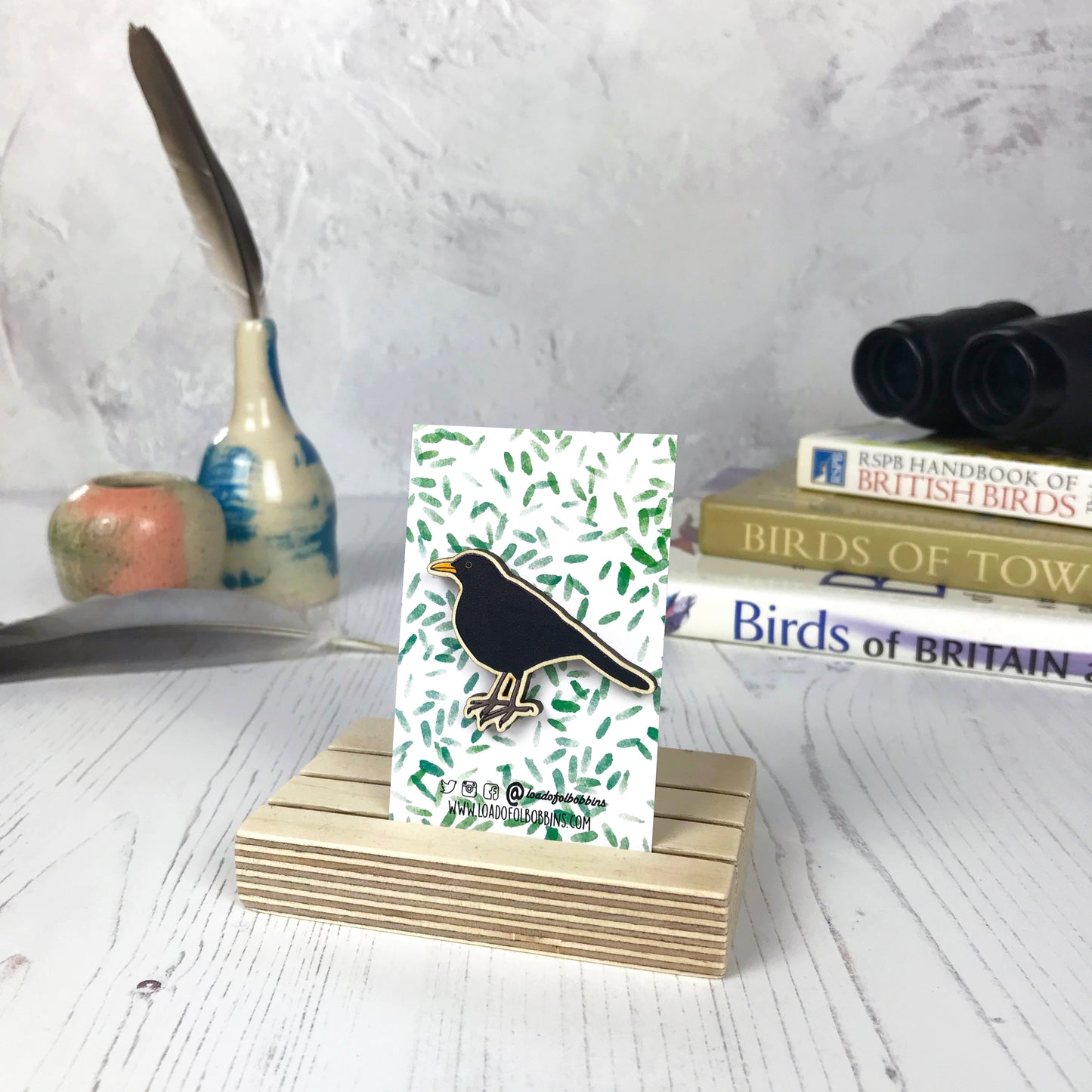 A laser cut wooden brooch showing an illustration of a male blackbird bird by loadofolbobbins. The brooch is mounted on a backing card with a repeat abstract watercolour leaf pattern. They are standing in a wooden stand and in the background you can see a pile of bird watching books and a pair of binoculars.