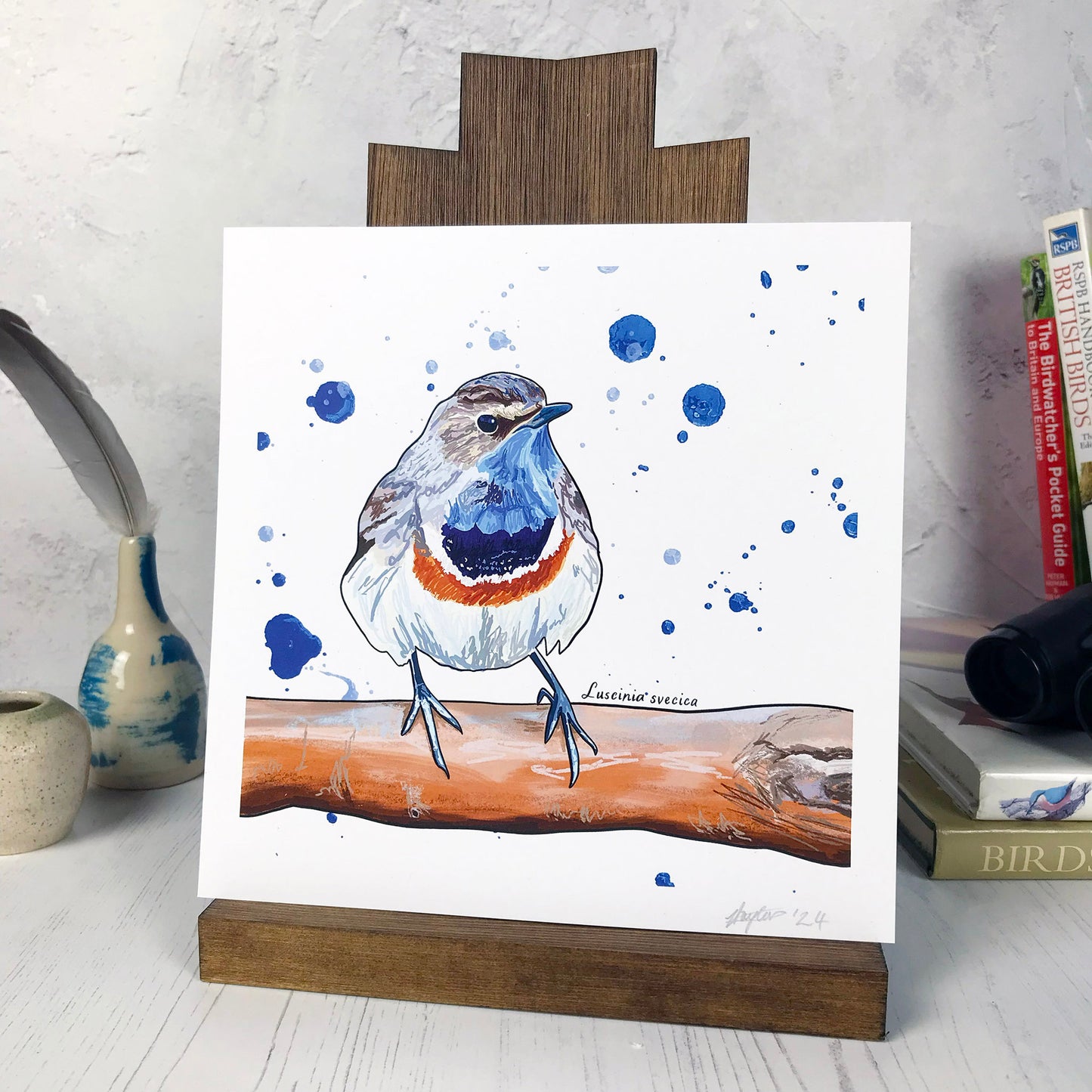 A photo of a medium sized 21 cm square art print made from an original digital illustration by Loadofolbobbins. It is of a bluethroat bird perching on a branch. It is similar in size to a robin but has a rich blue and orange throat pattern. It has a white belly and brownish head and wings. In the background are some rich deep blue paint drops. The illustration is made with energetic lines and a black outline. There is a 2.5 cm white border for framing.