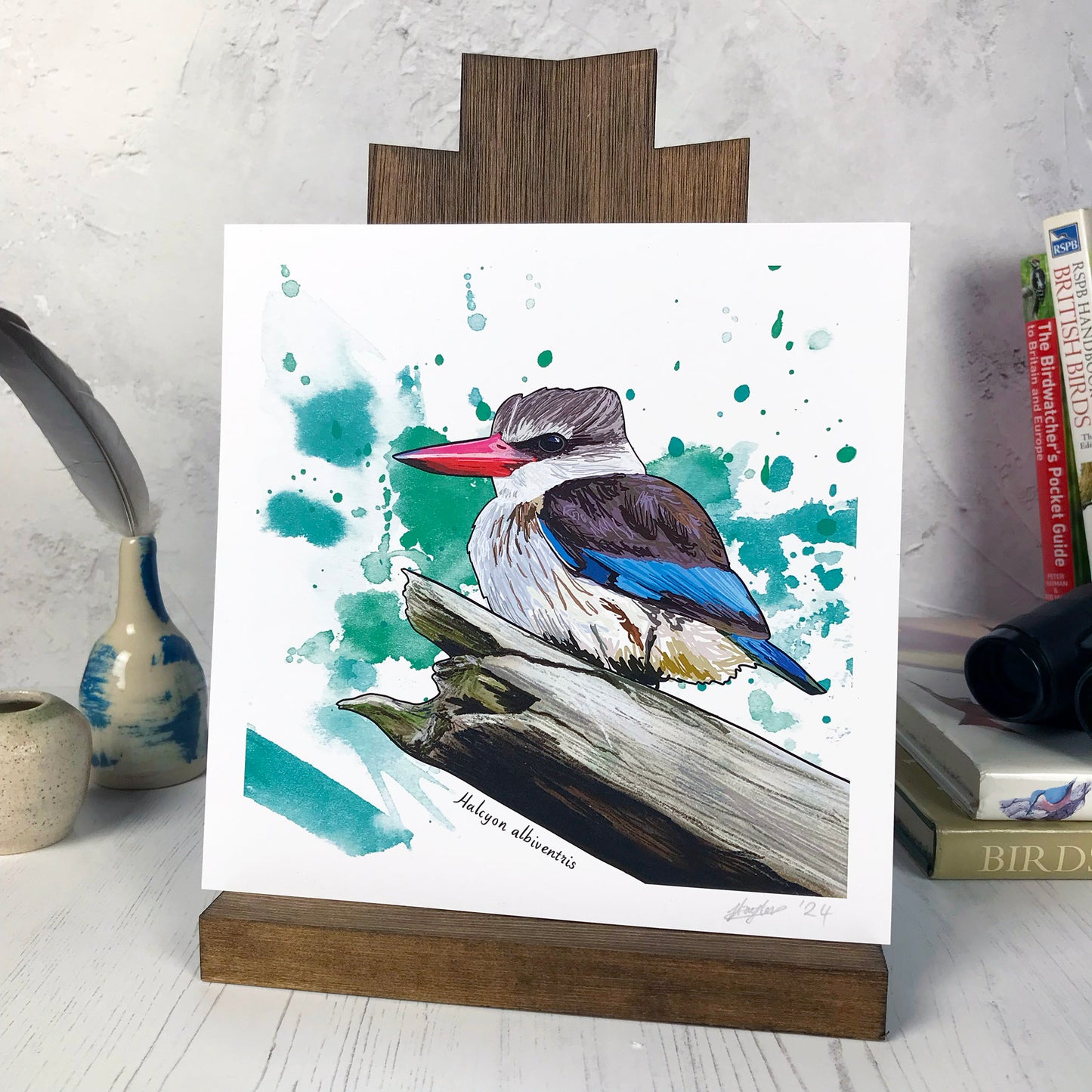 A photo of a medium sized 21 cm square art print made from an original digital illustration by Loadofolbobbins. It is of a brown hooded kingfisher perched on a large branch broken off at the end. It has a powerful fairly long sharp red beak. It's under feathers are a buff colour and it's wings and upper head are a a rich brown with vivid flashes of turquoise feathers in it's wings and tail feathers. The background is green inky splash background. There is a 2.5 cm white border for framing.