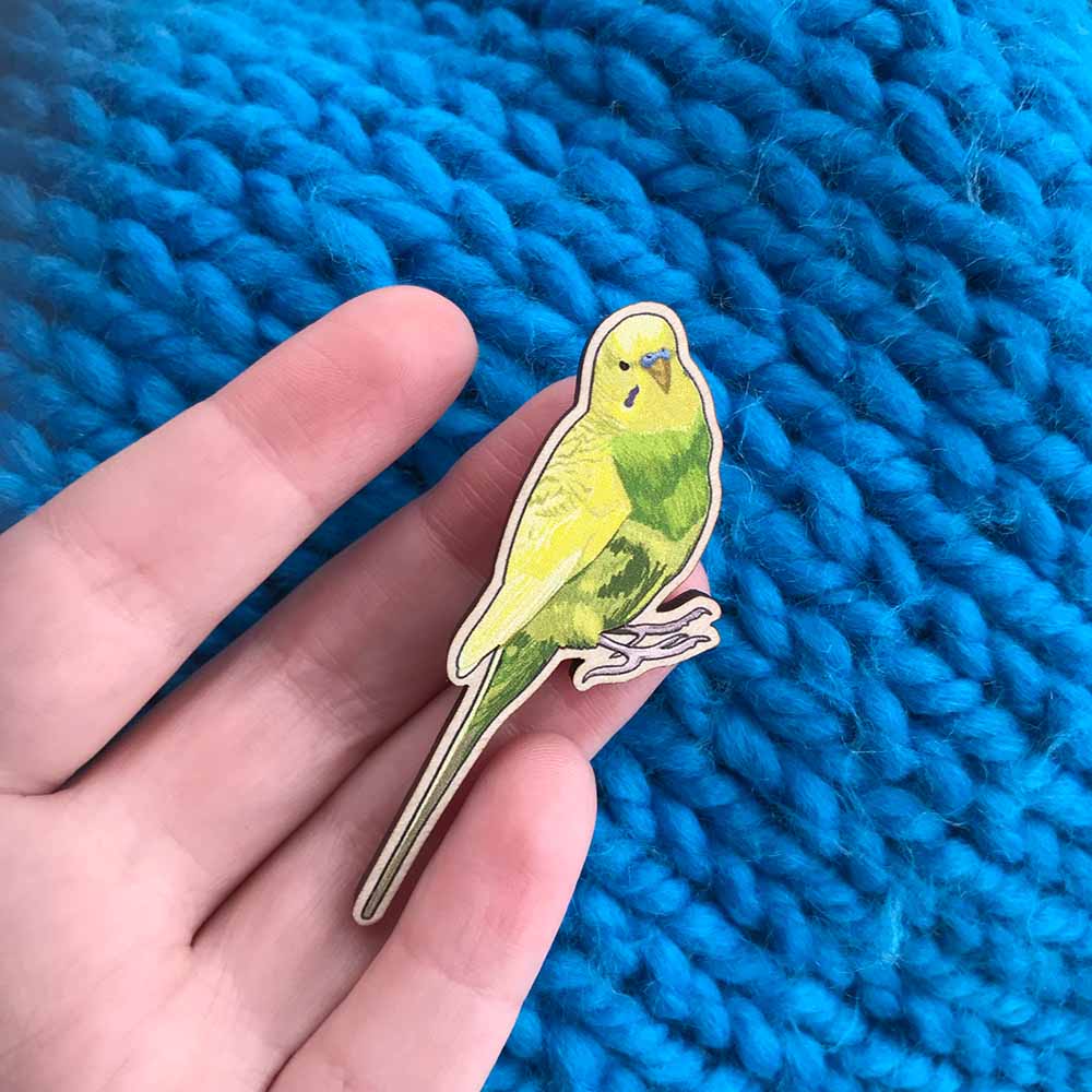 My hand, that of a white woman is holding a laser cut wooden brooch which shows an illustration by me Loadofolbobbins of a lime green and yellow budgie. The background of the image is a close up of a knitted turquoise cardigan.