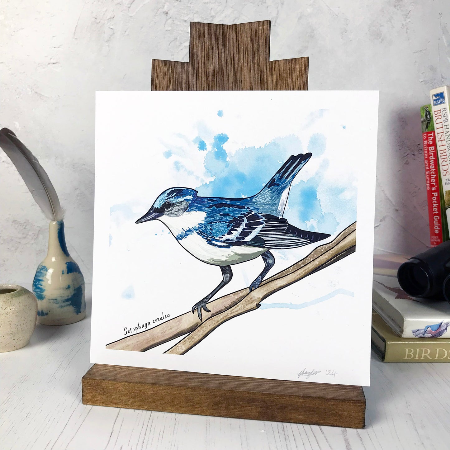 A photo of a medium sized 21 cm square art print made from an original digital illustration by Loadofolbobbins. It is of a cerulean warbler bird perching on a branch. It has been done with energetic line marks and has a light blue inky wash background. It is a small perky looking bird with deep blue feathers across it's back and white feathers underneath. There is a 2.5 cm white border for framing.