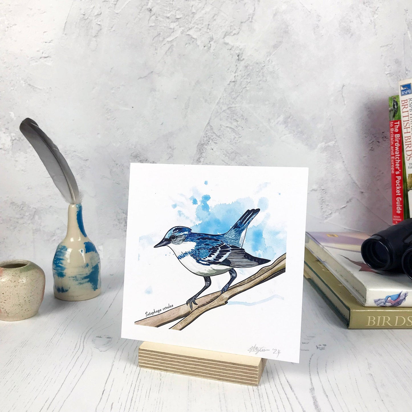 A photo of a small sized 14 cm square art print made from an original digital illustration by Loadofolbobbins. It is of a cerulean warbler bird perching on a branch. It has been done with energetic line marks and has a light blue inky wash background. It is a small perky looking bird with deep blue feathers across it's back and white feathers underneath. There is a 2.5 cm white border for framing.