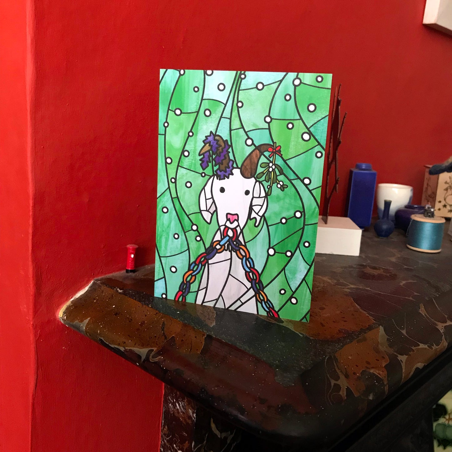A photo of a Christmas card on a mantel piece featuring an original illustration by Loadofolbobbins. The illustration shows a white goat with brown horns. The goat is eating part of a paper chain, its left horn is wrapped in purple tinsel and the right horn has mistletoe tied to it. The background is green with white snowy circles. The illustration is done in a stained glass style.