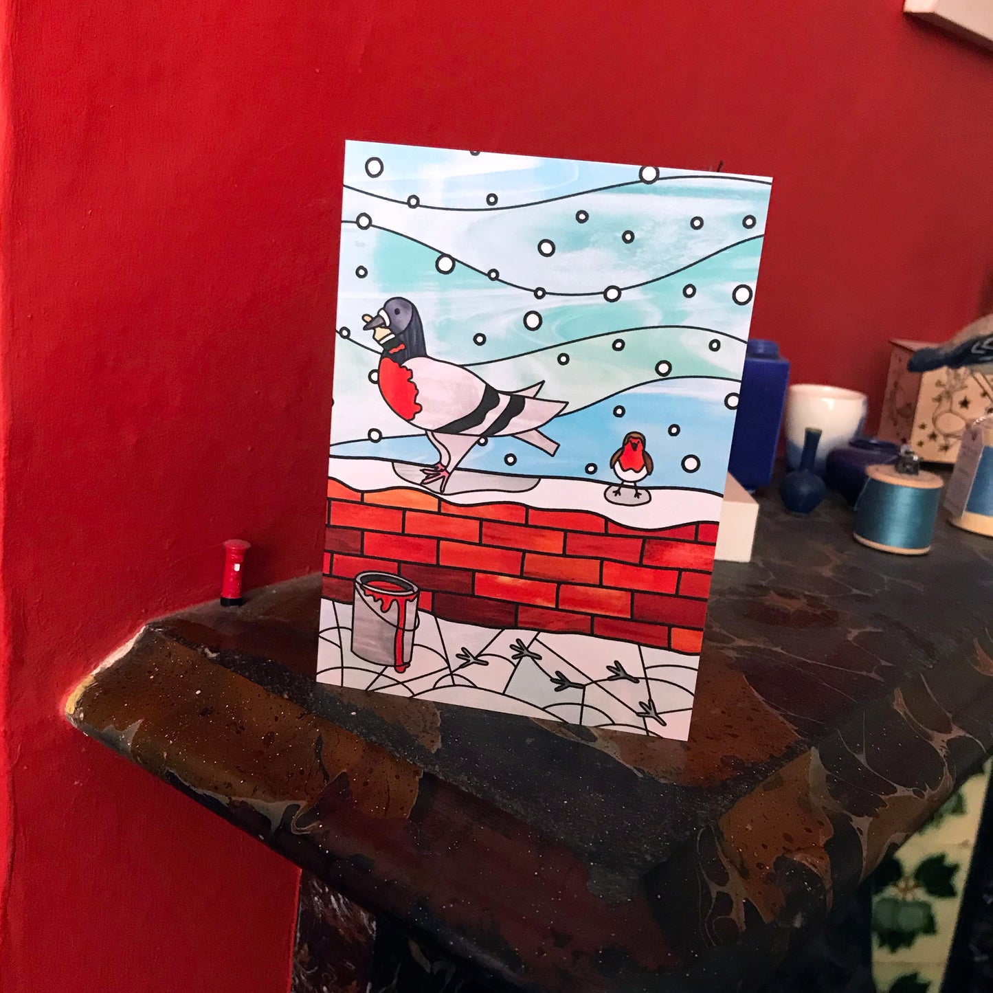 A photo of a Christmas card on a mantelpiece featuring an original illustration by Loadofolbobbins. The illustration shows a pigeon standing on a snow covered brick wall next to a robin. The pigeon has a paintbrush in it's beak and red paint covering it's chest to appear like a robin. In front of the brick wall is a can of opened red paint with pigeon prints in the snow leading to it. The background is light blue with white snowy circles. The illustration is done in a stained glass style.