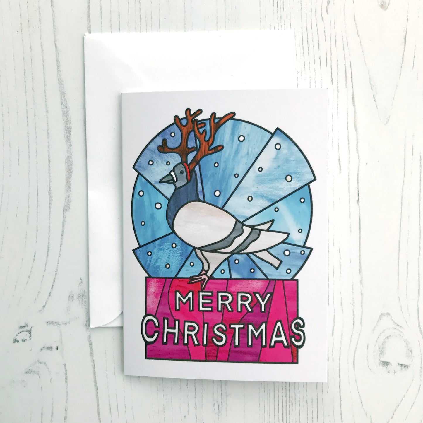 A photo of a Christmas card featuring an original illustration by Loadofolbobbins. The illustration shows a pigeon dressed up with reindeer antlers standing against a stylised circular blue snowy background. It is also standing atop white text that says Merry Christmas and has a hot pink background. The illustration is done in a stained glass style.