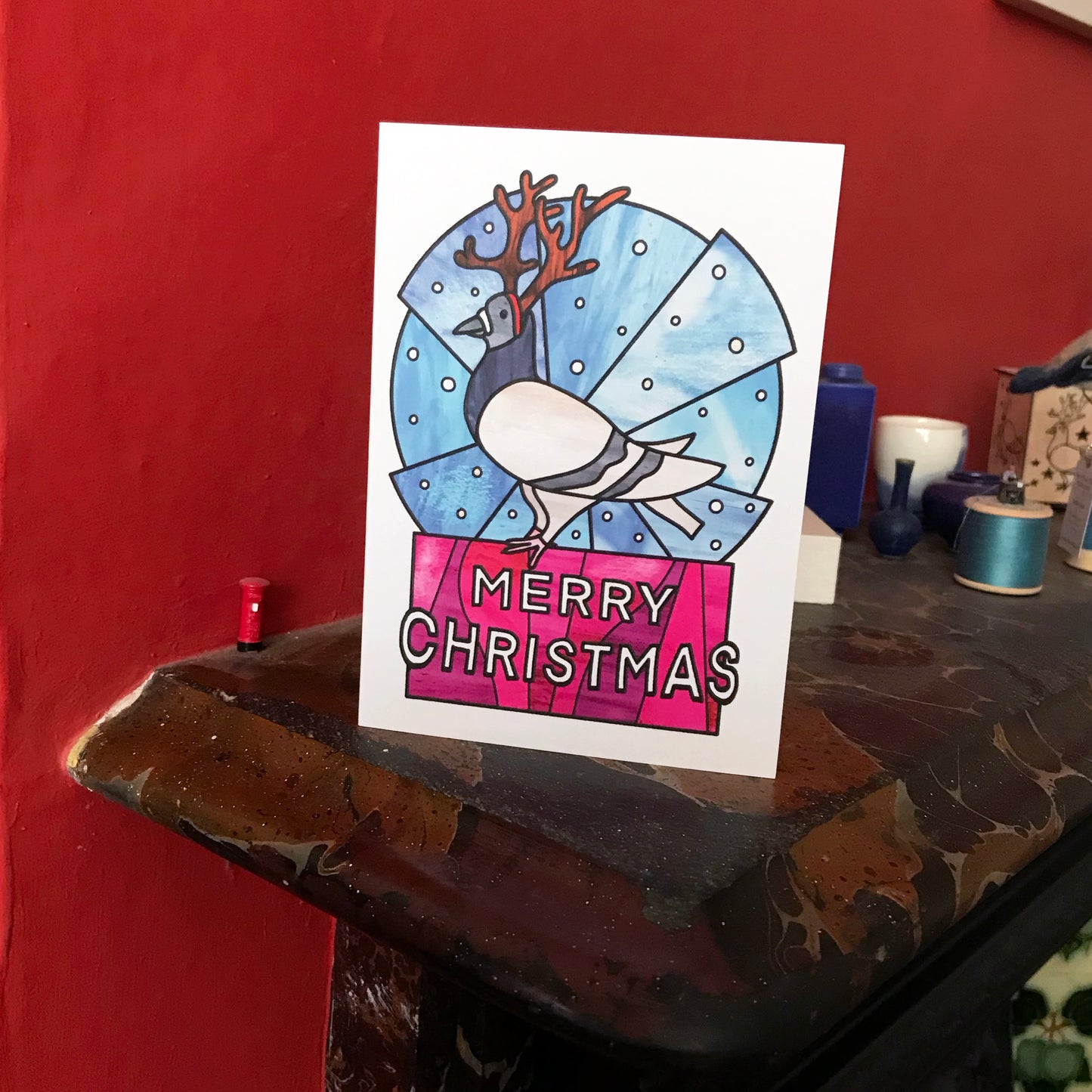 A photo of a Christmas card on a mantelpiece featuring an original illustration by Loadofolbobbins. The illustration shows a pigeon dressed up with reindeer antlers standing against a stylised circular blue snowy background. It is also standing atop white text that says Merry Christmas and has a hot pink background. The illustration is done in a stained glass style.