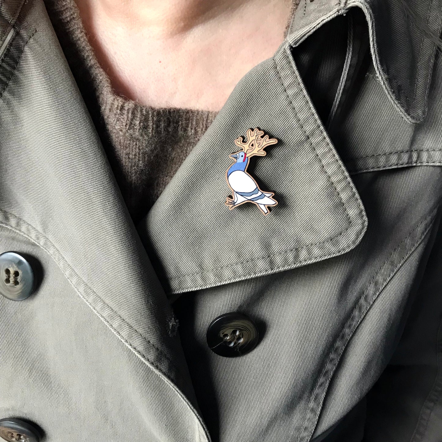 A laser cut wooden lapel pin showing an original illustration by Loadofolbobbins of a pigeon dressed up as a reindeer. The pigeon is wearing reindeer antlers. The style of the illustration is simplified with flat colours and a bold black outline. The pin is shown on the lapel of a jacket.