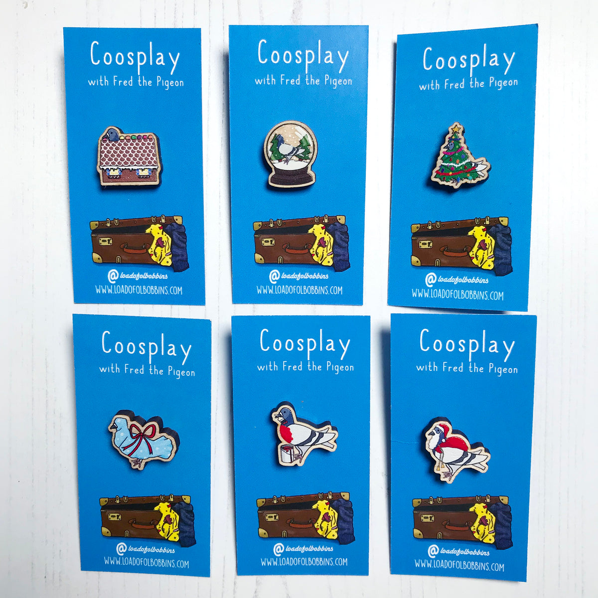 A group of six laser cut wooden 20mm lapel pins showing an original illustrations by Loadofolbobbins of pigeons in different Christmas themes. From left to right they show a gingerbread house, snow globe, christmas tree, santa claus, a robin and a Christmas gift. The style of the illustration is simplified with flat colours and a bold black outline. The pin is mounted on a blue backing card that says Coosplay with Fred the pigeon.