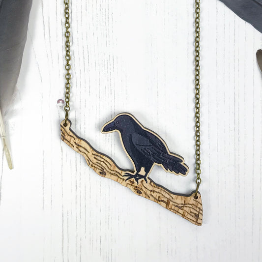 A laser cut wooden charm necklace showing an illustration of a crow perching on a branch by loadofolbobbins. There is pyrography texture detail on the branch.  The wooden necklace is hanging from an antique bronze cable chain.