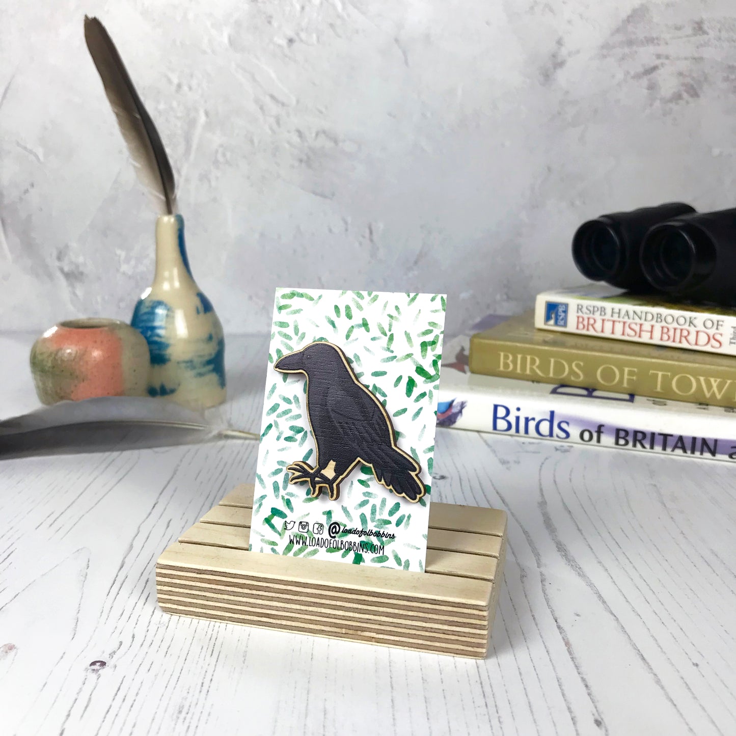 A laser cut wooden brooch showing an illustration of a crow bird by loadofolbobbins. The brooch is mounted on a backing card with a repeat abstract watercolour leaf pattern. They are standing in a wooden stand and in the background you can see a pile of bird watching books and a pair of binoculars.