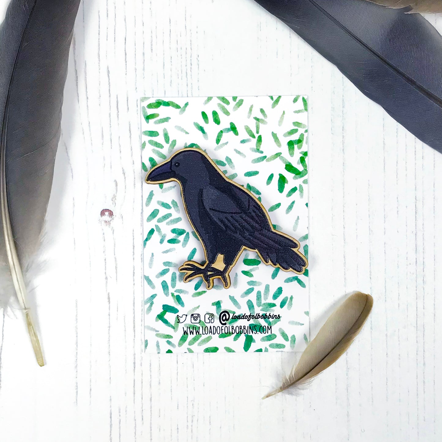 A laser cut wooden brooch showing an illustration by Loadofolbobbins of a crow. The brooch is mounted on a backing card with a repeat abstract watercolour leaf pattern.
