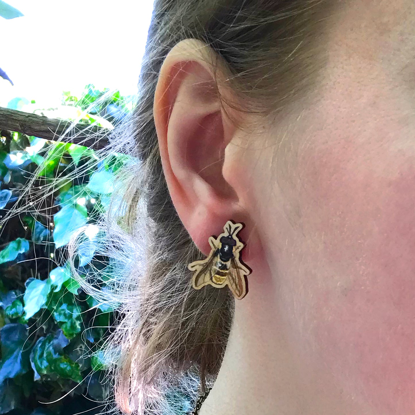 A close up photo of a white woman's ear with blondish brown hair worn up and standing against a wall of ivy. The main focus of the image is her ear which is wearing a small laser cut wooden stud earring of a golden hoverfly by Loadofolbobbins.