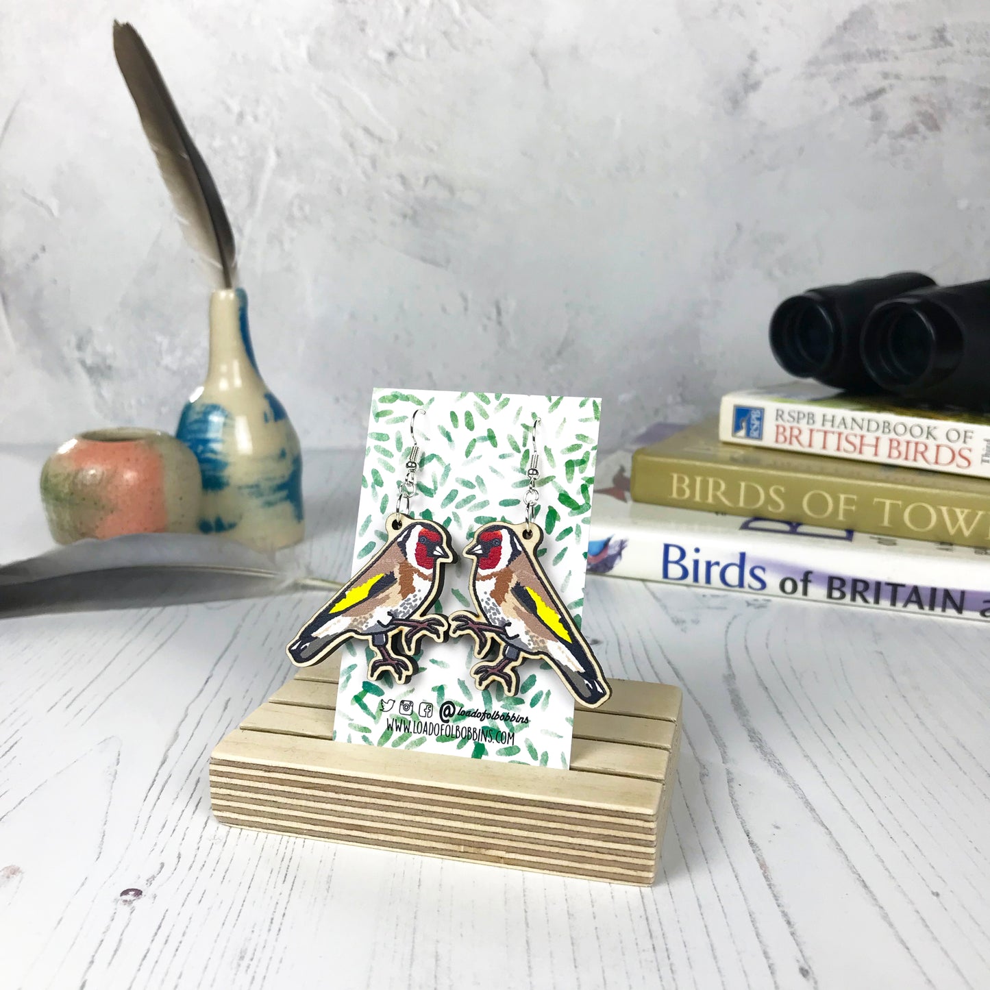 A pair of laser cut wooden dangle earrings showing an illustration of a goldfinch bird by loadofolbobbins. The earrings are mounted on a backing card with a repeat abstract watercolour leaf pattern. They are standing in a wooden stand and in the background you can see a pile of bird watching books and a pair of binoculars.