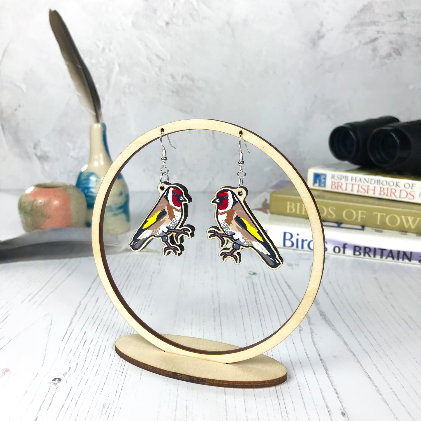 A pair of laser cut wooden dangle earrings showing an illustration of a goldfinch bird by loadofolbobbins. The earrings are hanging from a wooden circular ring display.