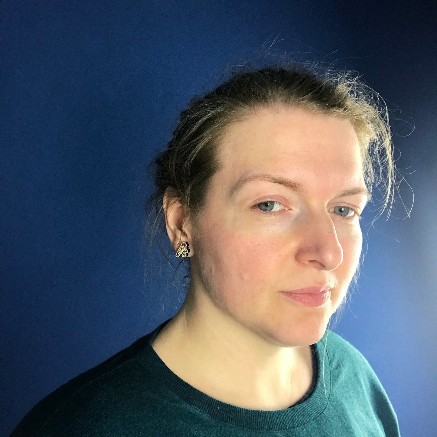 A neck and head close up photo of a white woman with blondish brown hair worn up and standing against a dark blue wall. Her face is slightly turned away from the camera so the main focus of the image is her ear which is wearing a small laser cut wooden stud earring of a goldfinch by Loadofolbobbins.