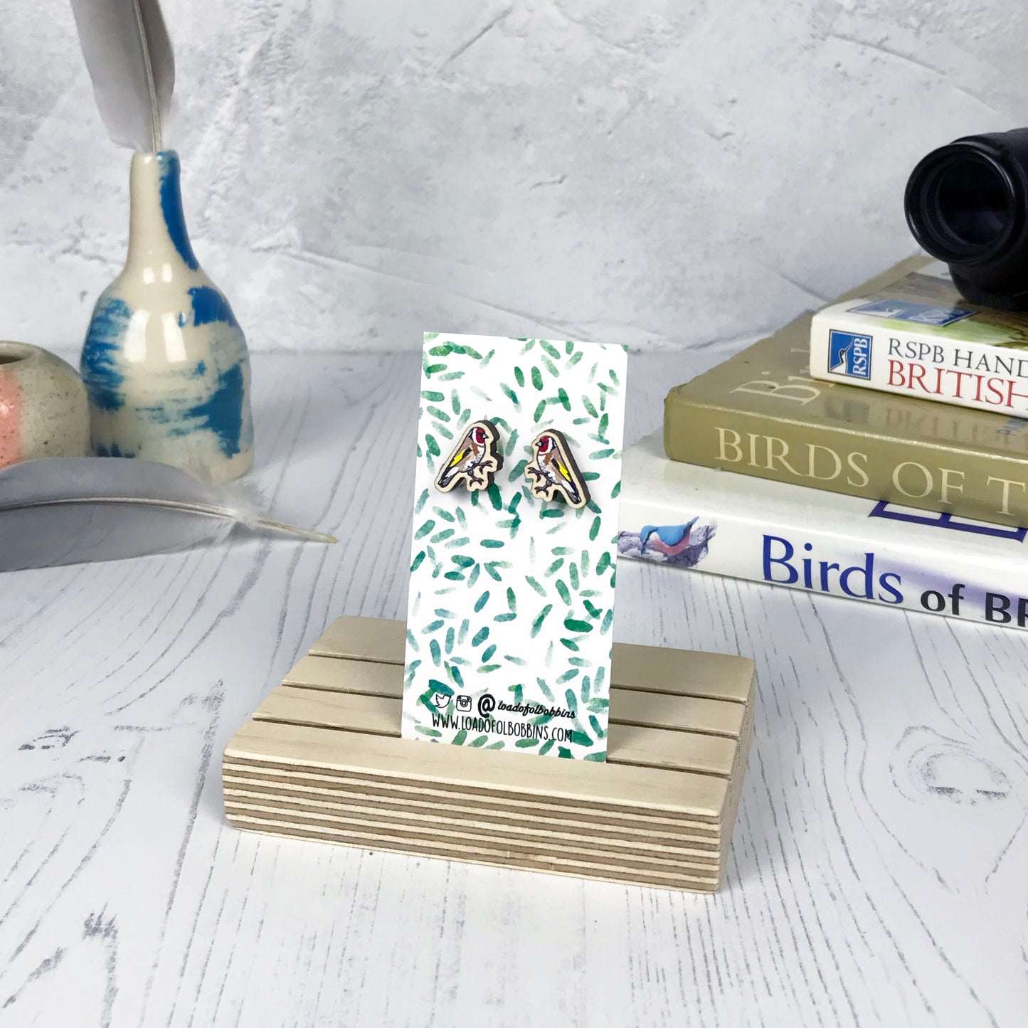 A pair of laser cut wooden earrings showing an illustration of a goldfinch bird by loadofolbobbins. The earrings are mounted on a mini backing card with a repeat abstract watercolour leaf pattern. They are standing in a wooden stand and in the background you can see a pile of bird watching books and a pair of binoculars.