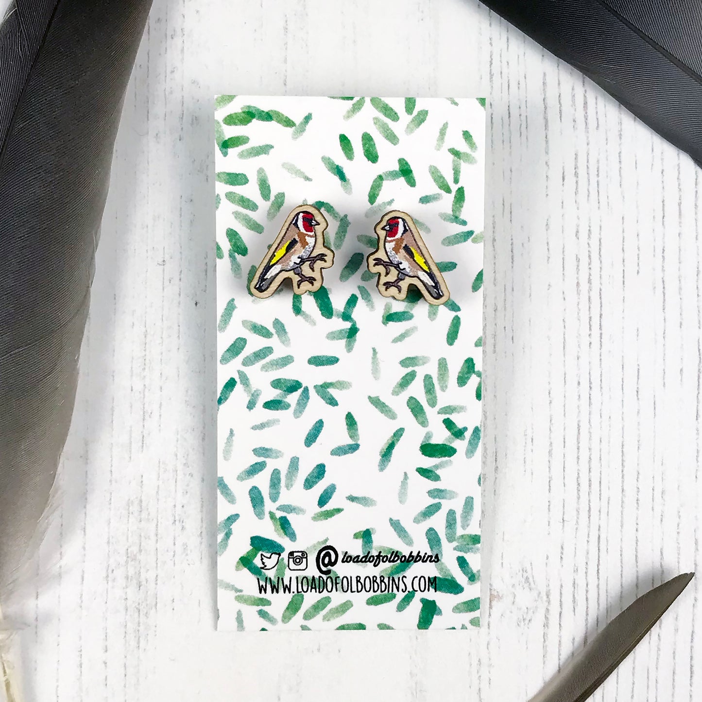 A pair of laser cut wooden earrings showing an illustration by Loadofolbobbins of a goldfinch. The earrings are mounted on a mini backing card with a repeat abstract watercolour leaf pattern.