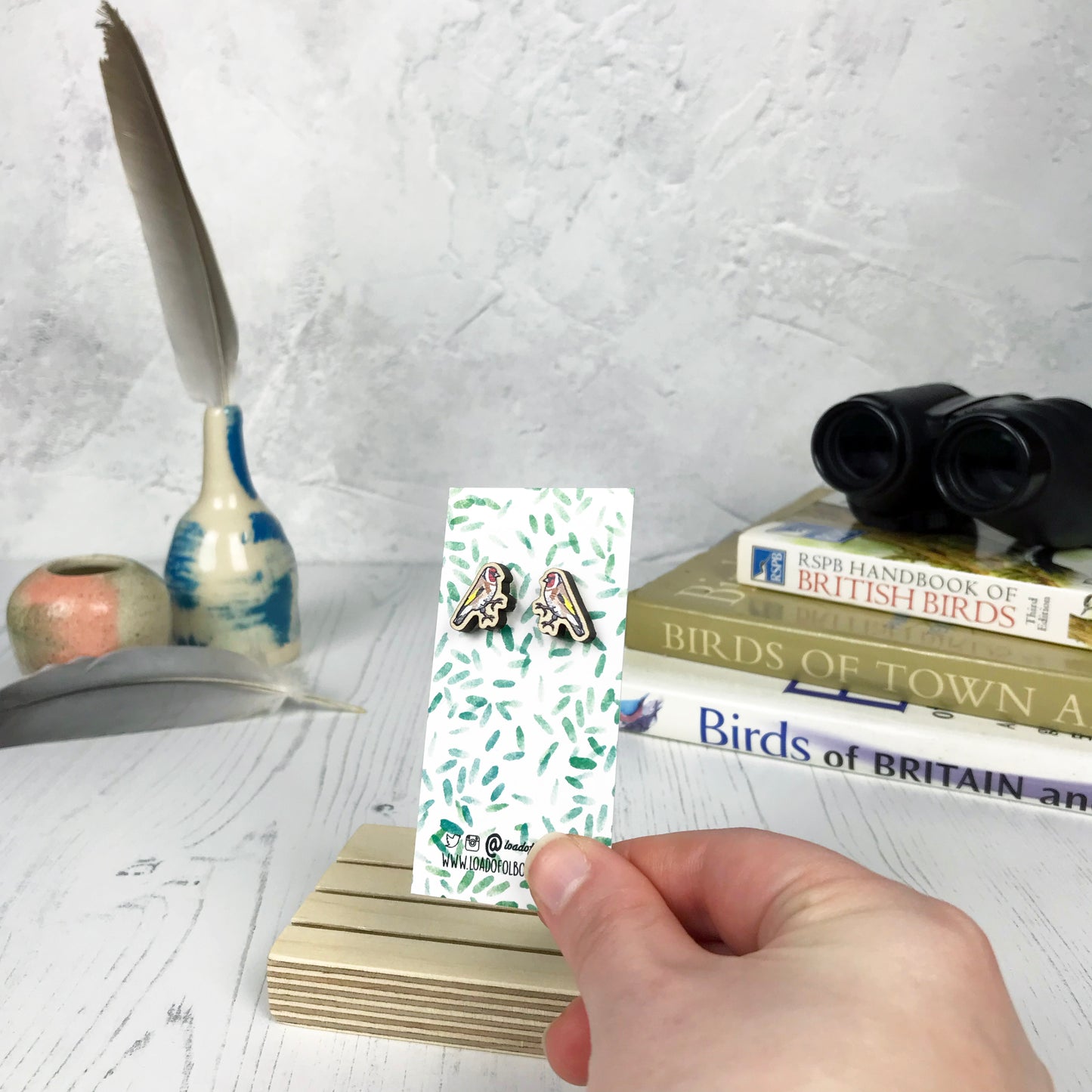 A pair of laser cut wooden earrings showing an illustration of a goldfinch bird by loadofolbobbins. The earrings are mounted on a mini backing card with a repeat abstract watercolour leaf pattern. They are being held by a hand for scale, which shows that each stud earring is slightly smaller than a thumbnail.
