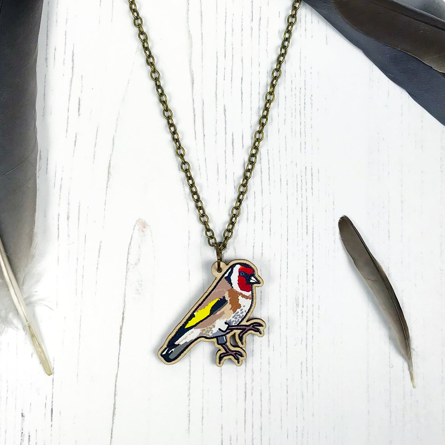A laser cut wooden charm necklace showing an illustration of a goldfinch bird by loadofolbobbins. The wooden charm is hanging from an antique bronze cable chain.