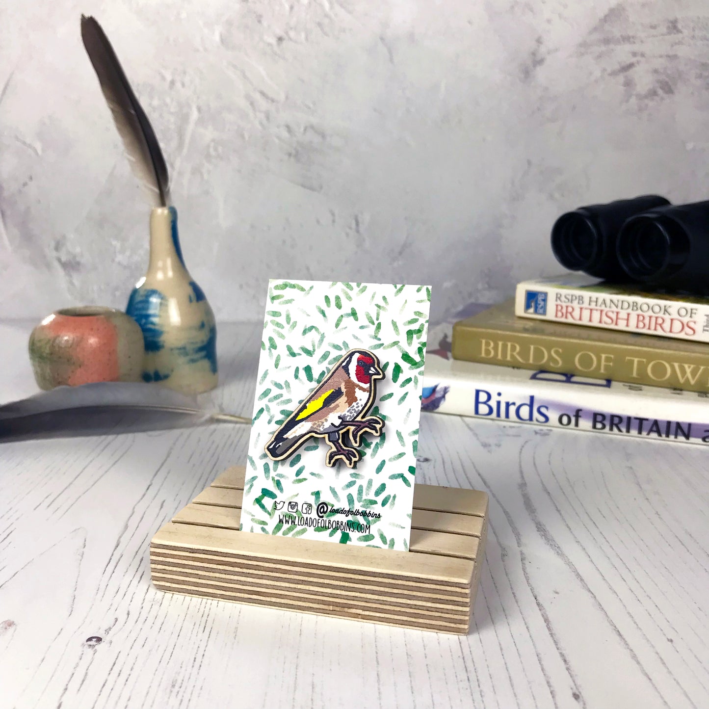 A laser cut wooden brooch showing an illustration of a goldfinch bird by loadofolbobbins. The brooch is mounted on a backing card with a repeat abstract watercolour leaf pattern. They are standing in a wooden stand and in the background you can see a pile of bird watching books and a pair of binoculars.