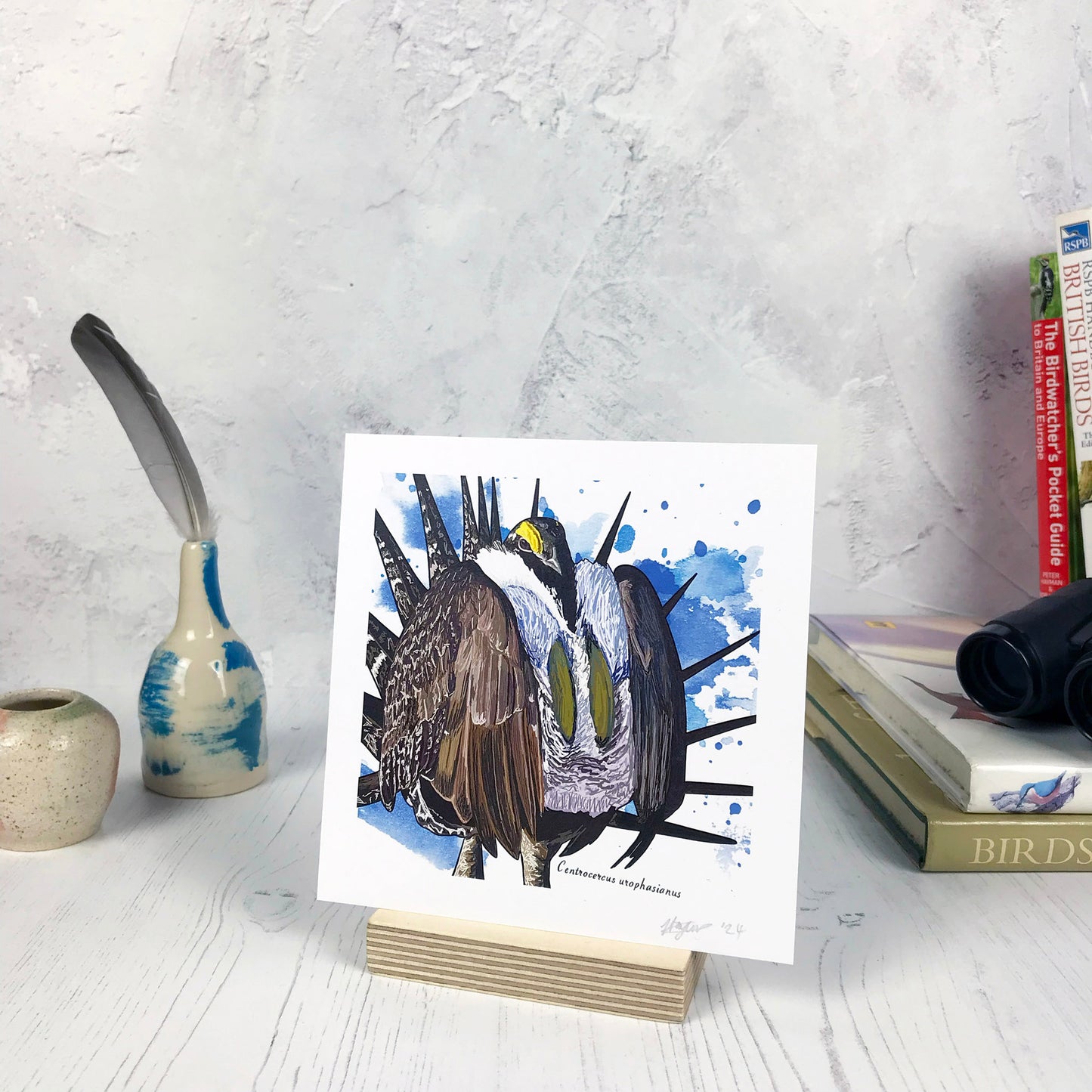 A photo of a small sized 14 cm square art print made from an original illustration by Loadofolbobbins. It is of a Greater sage grouse doing a mating display, it’s wings are puffed out like someone wearing too many coats and there are lots of spiky feathers pointed outwards like a sunburst. It has a very fluffy white inner chest feathers. It’s head is peeping out from amongst this with what looks like a bright yellow eyebrow. The background is blue inky splashes. There is a 2.5 cm white border for framing.