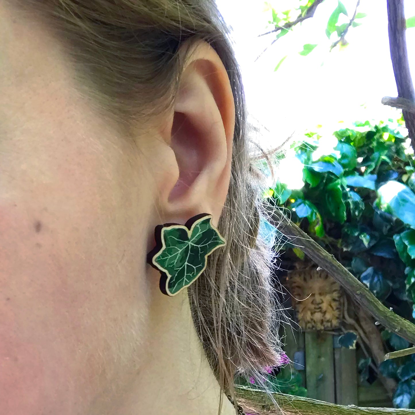 A close up photo of a white woman's ear with blondish brown hair worn up and standing against a wall of ivy. The main focus of the image is her ear which is wearing a small laser cut wooden stud earring of a dark green ivy leaf by Loadofolbobbins.