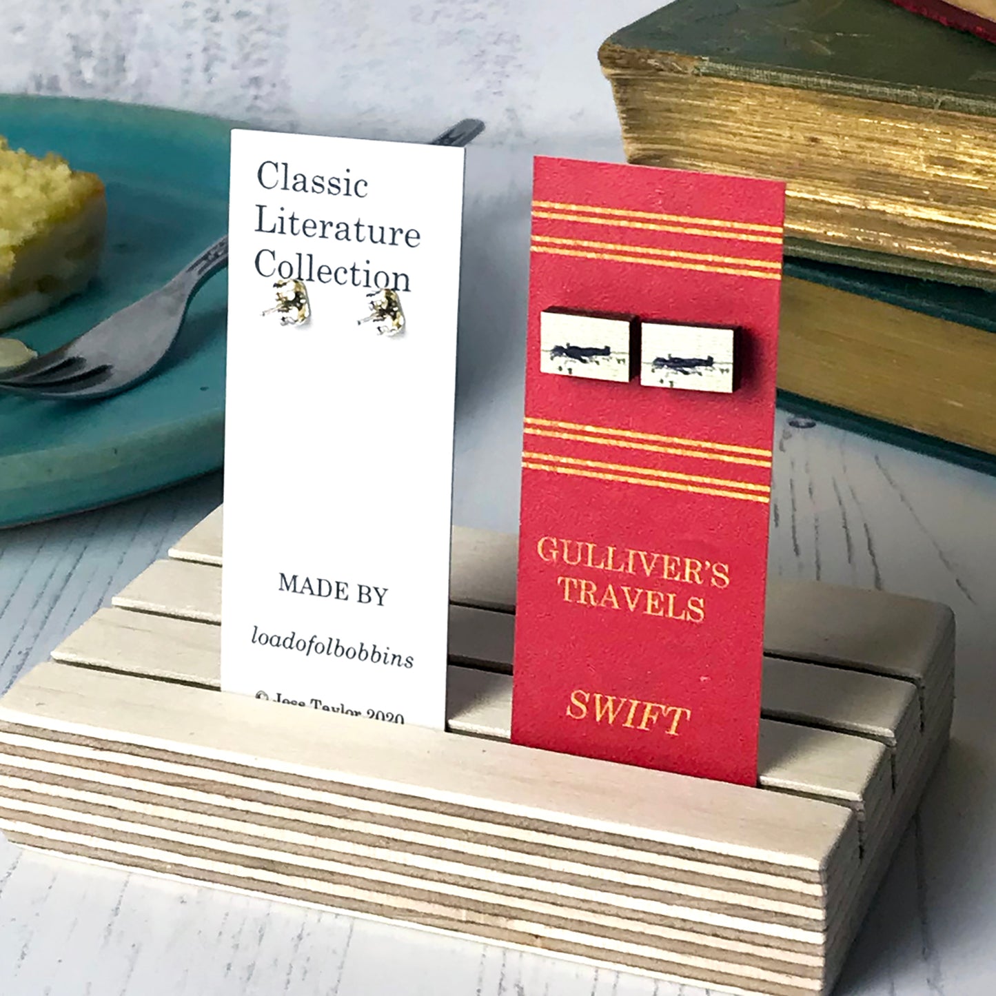 Laser cut wooden stud earrings by loadofolbobbins showing a silhouette scene inspired by the book Gulliver's Travels by Johnathan Swift. It shows Gulliver being tied to the ground by lilliputians. The scene is printed against original text from the novel. The earrings are mounted on a red backing card with gold lettering detail and made to look like the spine of a book.
