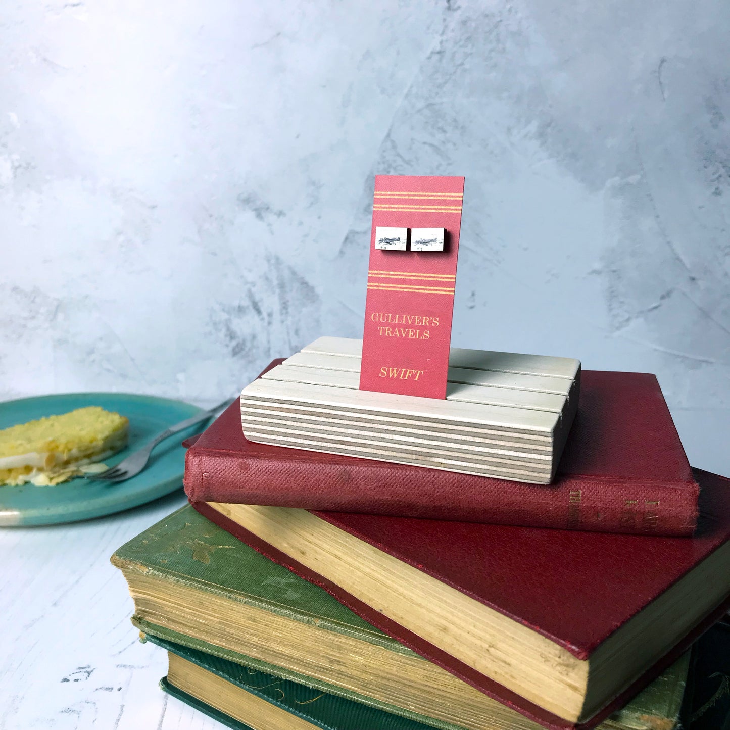 Laser cut wooden stud earrings by loadofolbobbins showing a silhouette scene inspired by the book Gulliver's Travels by Johnathan Swift. It shows Gulliver being tied to the ground by lilliputians. The scene is printed against original text from the novel. The earrings are mounted on a red backing card with gold lettering detail and made to look like the spine of a book.