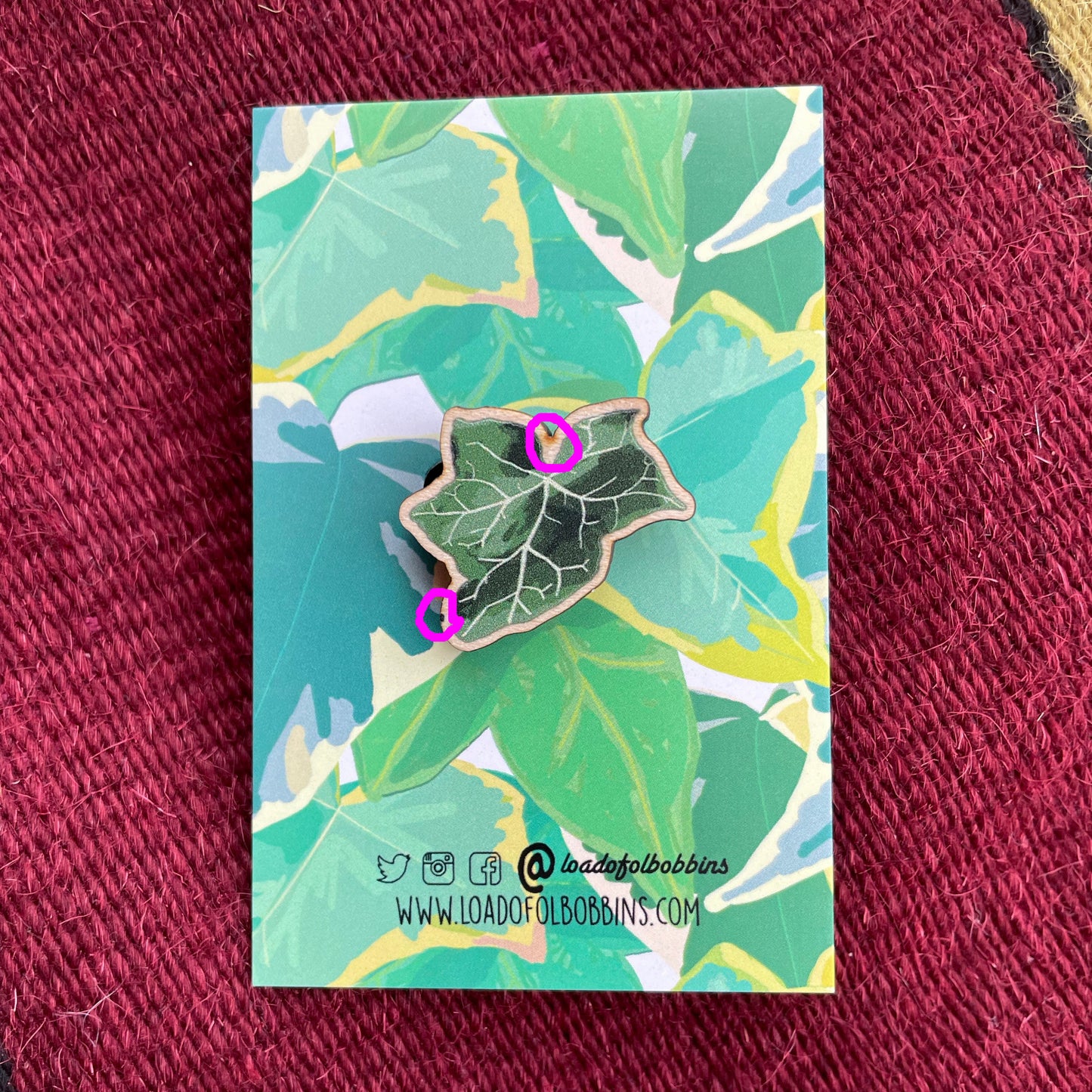 A small laser cut wooden brooch of a dark green ivy leaf. It is mounted on a backing card with a repeat pattern design of vibrant green ivy leaves.